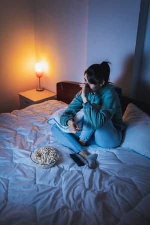 Téléchargez les photos : Belle jeune femme en pyjama assise au lit la nuit, triste en regardant un film dramatique à la télévision, étreignant un oreiller et pleurant - en image libre de droit