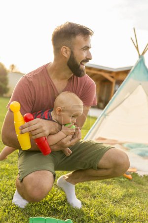Foto de Joven padre divirtiéndose jugando con su bebé, acampando en el patio trasero en un día soleado de verano, sentado en el césped cerca de la tienda de campaña y sosteniendo al hijo en su regazo - Imagen libre de derechos