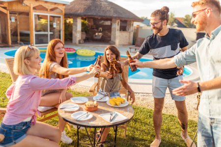Foto de Grupo de alegres jóvenes amigos divirtiéndose en la fiesta de barbacoa en el patio trasero junto a la piscina, haciendo un brindis con botellas de cerveza y disfrutando de días soleados de verano al aire libre - Imagen libre de derechos