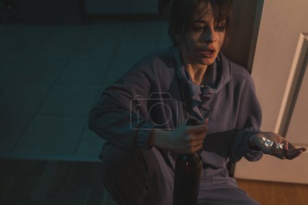 Foto de Mujer joven desesperada sentada en el suelo de la cocina en la oscuridad borracha, sosteniendo una botella de vino y un puñado de pastillas; abuso de sustancias, depresión y concepto de suicidio - Imagen libre de derechos