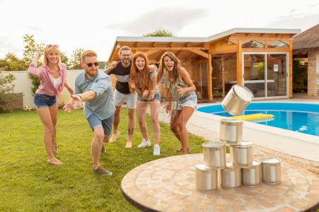 Gruppe fröhlicher junger Freunde, die Spaß daran haben, auf einer sommerlichen Outdoor-Party am Pool Blechdosen umzuwerfen, indem sie ein Ballspiel spielen