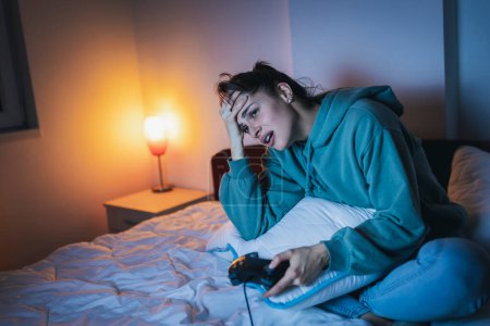 Photo for Beautiful young woman wearing pajamas having fun at home, playing video games in bed at night, disappointed after losing the game - Royalty Free Image