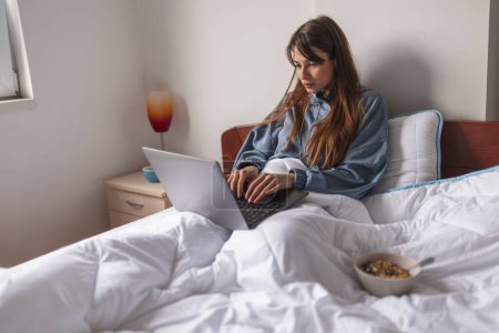 Téléchargez les photos : Jeune femme portant un pyjama assis au lit le matin, travaillant à la maison à l'aide d'un ordinateur portable et prenant le petit déjeuner au lit, mangeant des céréales - en image libre de droit
