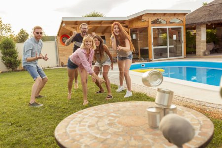 Gruppe fröhlicher junger Freunde, die Spaß daran haben, auf einer sommerlichen Outdoor-Party am Pool Blechdosen umzuwerfen, indem sie ein Ballspiel spielen