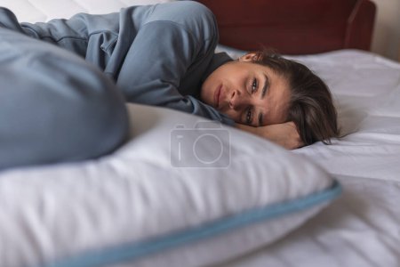 Foto de Mujer joven acostada acurrucada en la cama con dolor de estómago durante su período; mujer solitaria deprimida acostada en la cama por la mañana - Imagen libre de derechos