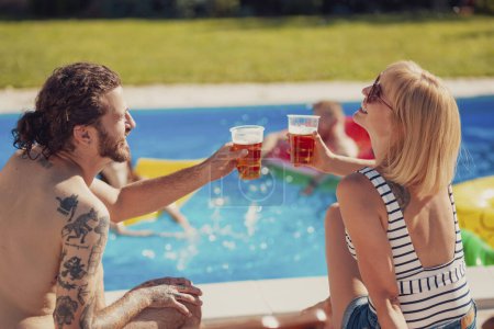 Hermosa pareja joven sentada junto a la piscina, tomando el sol y haciendo un brindis con vasos de cerveza mientras se relaja al aire libre en el soleado día de verano.
