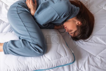 Top view of a young woman lying curled up in bed in pain having period cramps; depressed woman lying in bed in the morning