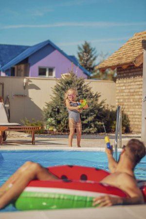 Téléchargez les photos : Beau père et fille joyeux jouant à la piscine et s'amusant à l'extérieur par une chaude journée ensoleillée d'été, éclaboussant l'eau avec des pistolets à eau - en image libre de droit
