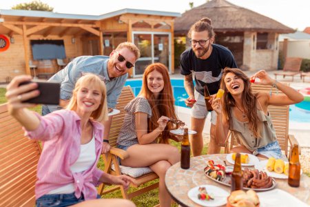 Foto de Grupo de amigos teniendo una fiesta de barbacoa junto a la piscina del patio trasero, divirtiéndose tomando selfies mientras se reúnen alrededor de la mesa - Imagen libre de derechos