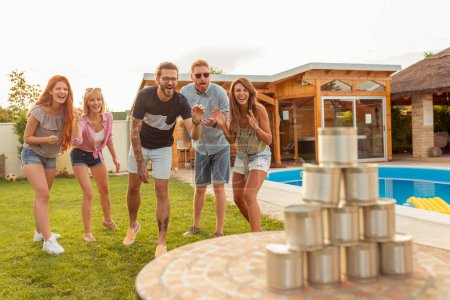 Photo for Group of cheerful young friends having fun playing the knock down tin cans by throwing a ball game while at poolside summertime outdoor party - Royalty Free Image
