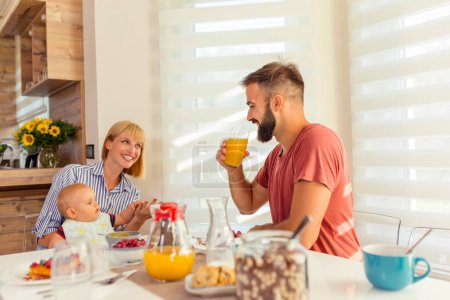 Foto de Los padres jóvenes se divierten desayunando y alimentando a su pequeño bebé, disfrutando pasar las mañanas de fin de semana juntos en casa - Imagen libre de derechos