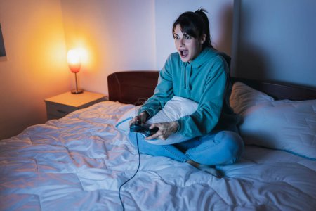 Téléchargez les photos : Belle jeune femme en pyjama s'amusant à jouer à des jeux vidéo au lit la nuit - en image libre de droit