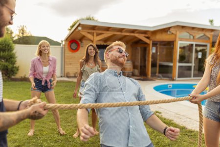 Téléchargez les photos : Groupe de jeunes amis joyeux s'amusant à la fête en plein air de l'été près de la piscine, participant à un concours de danse limbo, passant sous la corde tout en dansant - en image libre de droit
