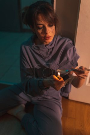 Foto de Un drogadicto sentado en el suelo en la oscuridad, preparando la siguiente dosis de cocaína intravenosa, usando un encendedor y una cuchara; concepto de adicción a las drogas y abuso de sustancias - Imagen libre de derechos