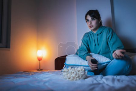 Photo for Beautiful young woman wearing pajamas sitting in bed at night, eating popcorn and watching a movie on TV - Royalty Free Image