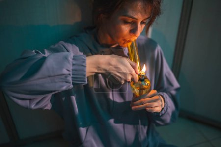 Photo for Depressed young woman sitting on the floor at dark, smoking pot using bong; woman vaping marijuana with bong - Royalty Free Image