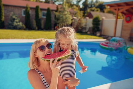 Téléchargez les photos : Belle mère et fille s'amusent à passer une chaude journée ensoleillée en plein air, à manger une tranche de pastèque près de la piscine - en image libre de droit