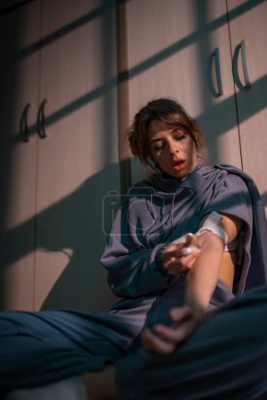 Foto de Mujer sentada en el suelo en la oscuridad inyectándose heroína; mujer drogadicta inyectándose dosis de heroína por vía intravenosa - Imagen libre de derechos