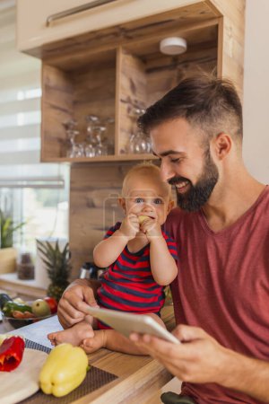 Photo for Father having fun spending time at home with his baby boy, sitting at kitchen counter, eating vegetables and watching cartoons - Royalty Free Image