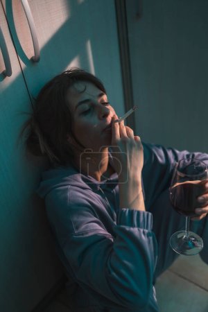 Foto de Mujer deprimida sentada en el suelo en la oscuridad, bebiendo vino y fumando cigarrillo, molesta y desesperada - Imagen libre de derechos