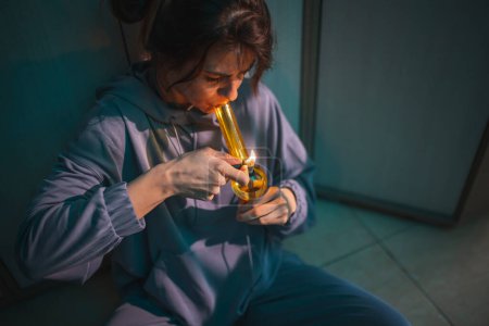 Téléchargez les photos : Jeune femme déprimée assise sur le sol à la tombée de la nuit, fumant de l'herbe avec du bong ; femme vaporisant de la marijuana avec du bong - en image libre de droit
