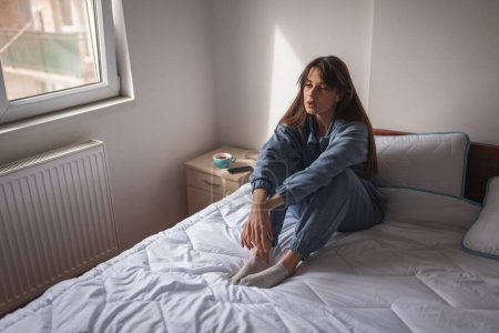 Foto de Mujer joven con pijama sentado en la cama por la mañana, pensativo y serio, que tiene problemas con la depresión y la soledad - Imagen libre de derechos