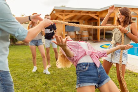Foto de Grupo de alegres jóvenes amigos divirtiéndose en la fiesta al aire libre de verano junto a la piscina, participando en el concurso de baile del limbo, pasando por debajo de la cuerda mientras bailan - Imagen libre de derechos