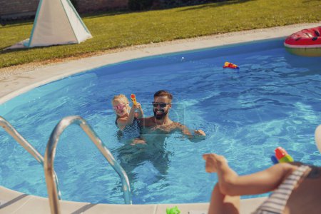 Téléchargez les photos : Les jeunes parents s'amusent à jouer avec leur fille dans la piscine, éclaboussant l'eau avec des pistolets à eau, profitant d'une chaude journée d'été ensoleillée à l'extérieur et se relaxant pendant les vacances d'été - en image libre de droit
