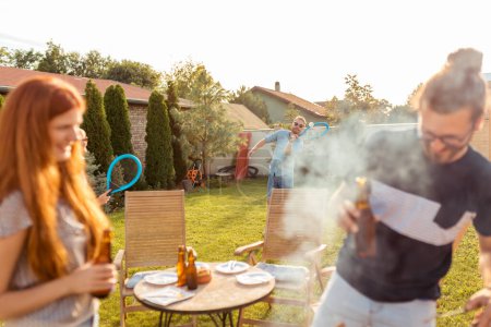 Téléchargez les photos : Groupe de jeunes amis joyeux ayant une fête barbecue arrière-cour, griller de la viande, boire de la bière, jouer au badminton et se détendre par une journée d'été ensoleillée en plein air - en image libre de droit