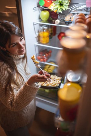 Téléchargez les photos : Vue en angle élevé d'une belle jeune femme debout à côté d'une porte de réfrigérateur ouverte la nuit, mangeant des pâtes et choisissant la nourriture - en image libre de droit