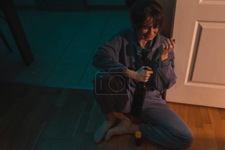 Photo for High angle view of depressed young woman sitting on the kitchen floor in the dark crying, drinking wine and pills; substance abuse, depression and suicide concept - Royalty Free Image