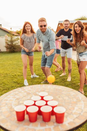 Foto de Grupo de alegres jóvenes amigos que se divierten mientras juegan al pong de cerveza en la fiesta de verano del patio trasero, animándose mutuamente - Imagen libre de derechos