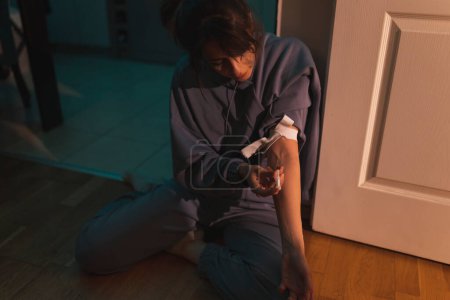 Téléchargez les photos : Femme assise sur le sol dans le noir tirant avec de l'héroïne ; toxicomane s'injectant elle-même de l'héroïne par voie intraveineuse ; toxicomanie et abus de substances concéder - en image libre de droit