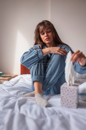 Photo for Sad young woman wearing pajamas, sitting on bed in the morning, using paper tissues for wiping tears and nose after crying - Royalty Free Image