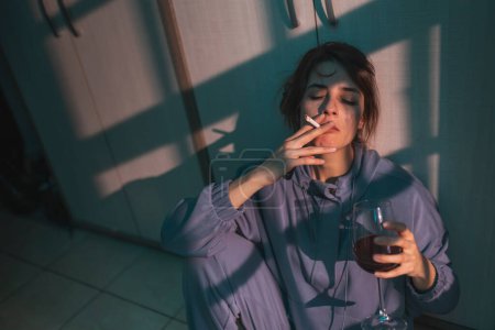 Téléchargez les photos : Jeune femme déprimée assise par terre dans le noir, buvant du vin et fumant de la cigarette, bouleversée et désespérée - en image libre de droit