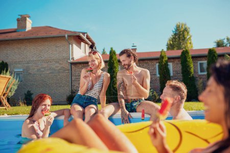 Photo for Group of young friends having fun at summertime swimming pool party, sitting by the pool, floating on a swimming floats and eating watermelon popsicles - Royalty Free Image