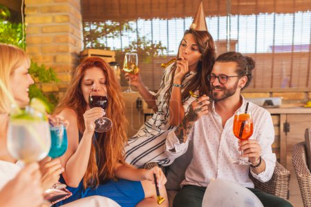Foto de Grupo de alegres jóvenes amigos divirtiéndose bebiendo cócteles y soplando silbatos de fiesta en una fiesta de cumpleaños - Imagen libre de derechos