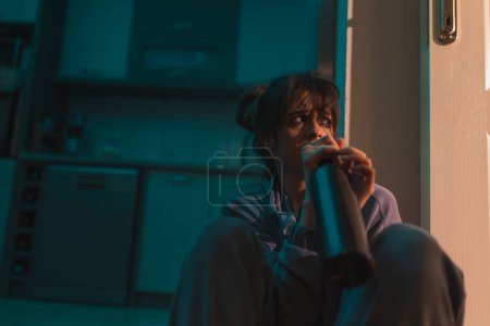 Photo for Depressed drunk woman sitting on the floor in the dark with smeared makeup, drinking wine from a bottle and crying - Royalty Free Image