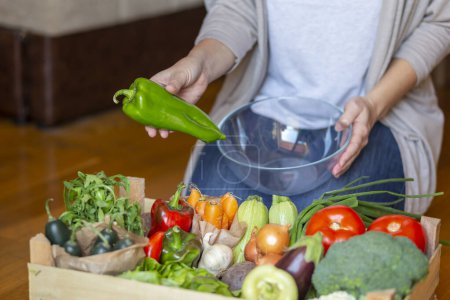 Foto de Mujer sosteniendo un tazón y seleccionando verduras orgánicas frescas para la preparación del almuerzo de una caja de madera de entrega de alimentos - Imagen libre de derechos