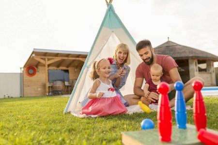 Foto de Feliz familia divirtiéndose pasando el soleado día de verano acampando en el patio trasero, los padres jugando bolos con sus hijos pequeños, lanzando una pelota y derribando los alfileres - Imagen libre de derechos