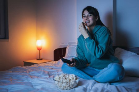 Téléchargez les photos : Jeune femme en pyjama assis au lit, manger du pop-corn et rire en regardant un film drôle à la télévision, se détendre à la maison tard dans la nuit - en image libre de droit