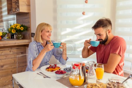 Téléchargez les photos : Beau jeune couple amoureux assis à la table de la salle à manger, boire le café du matin et prendre le petit déjeuner, passer le week-end matin ensemble à la maison - en image libre de droit