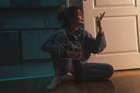 Téléchargez les photos : Femme ivre déprimée assise sur le sol dans le noir avec un maquillage enduit, buvant du vin dans une bouteille et pleurant - en image libre de droit
