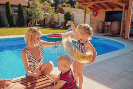 Téléchargez les photos : Jeune mère jouant avec les petits enfants près de la piscine par une chaude journée ensoleillée d'été, petite fille essayant sur anneau de natation pendant que le bébé garçon mange pastèque - en image libre de droit