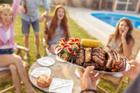 Téléchargez les photos : Groupe de jeunes amis joyeux réunis autour de la table, boire de la bière et s'amuser à une fête barbecue dans la piscine, hôte apportant assiette de viande et légumes grillés - en image libre de droit