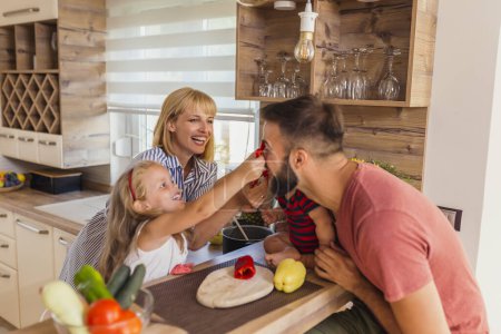 Foto de Familia feliz divirtiéndose mientras cocinan en la cocina, madre e hija sosteniendo trozos de pimiento como gafas y haciendo caras divertidas - Imagen libre de derechos