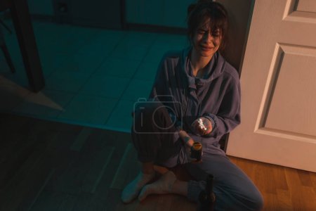 Foto de Vista de ángulo alto de la mujer joven deprimida sentada en el suelo de la cocina en la oscuridad llorando, sosteniendo una botella de vino y un puñado de pastillas, pensando en el suicidio - Imagen libre de derechos