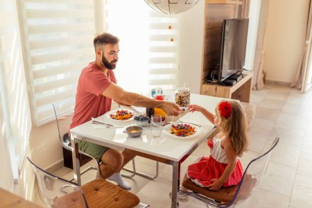 Foto de Padre joven y hermosa hija pequeña sentados en la mesa del comedor, desayunando en casa y disfrutando de su tiempo juntos - Imagen libre de derechos