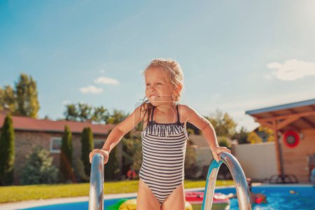 Foto de Hermosa niña con traje de baño de pie en la escalera de la piscina, salir de la piscina y divertirse al aire libre con su padre en un día de verano soleado caliente - Imagen libre de derechos