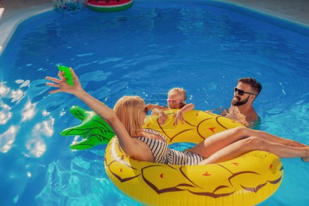 Téléchargez les photos : Les jeunes parents s'amusent à jouer avec leur fille dans la piscine, profiter d'une chaude journée ensoleillée en plein air et se détendre pendant les vacances d'été - en image libre de droit